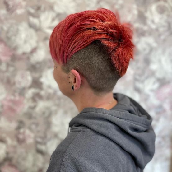 Eine Person mit rotem Haar, das in einem Irokesenschnitt gestylt ist, steht mit dem Rücken zur Kamera und trägt eine graue Kapuzenjacke.