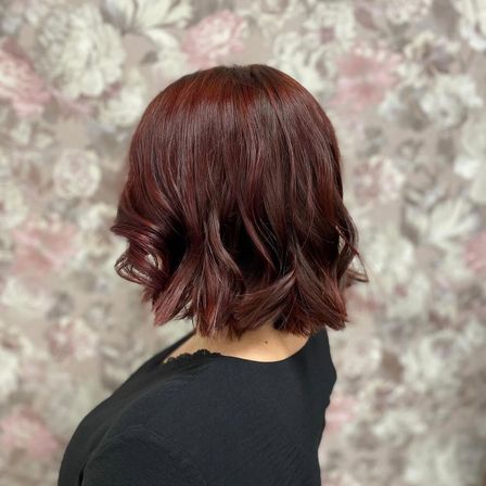 Eine Frau mit kurzen, gewellten, burgunderroten Haaren in einem Bob-Schnitt, trägt ein schwarzes Oberteil und steht vor einem floralen Hintergrund.