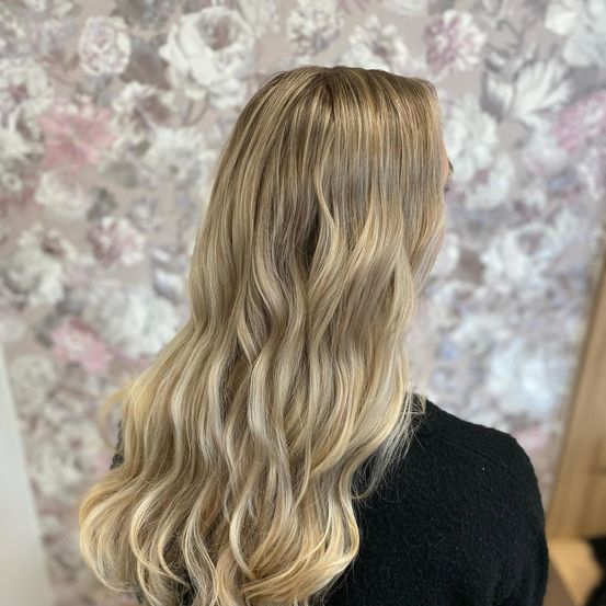 Eine Frau mit langen, gewellten blonden Haaren steht vor einer floralen Tapete. Sie trägt ein schwarzes Oberteil.