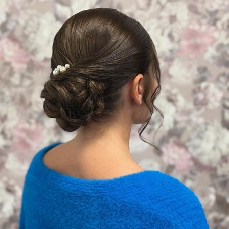 Eine Frau mit braunen Haaren, die in einem Dutt mit einer Perlenhaarnadel gestylt sind. Sie trägt einen blauen Pullover.