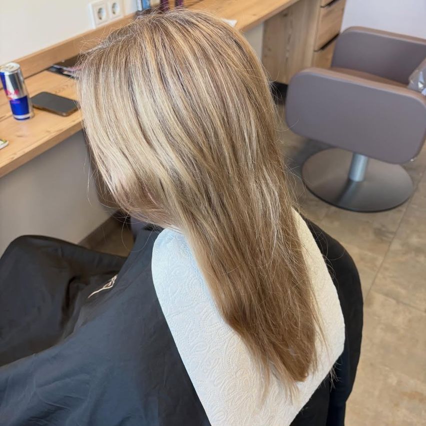 Eine Frau mit langen blonden Haaren sitzt in einem Salonstuhl mit einem weißen Umhang. Dahinter steht ein Holzschreibtisch mit einem Telefon, einer Dose und einer Steckdose.