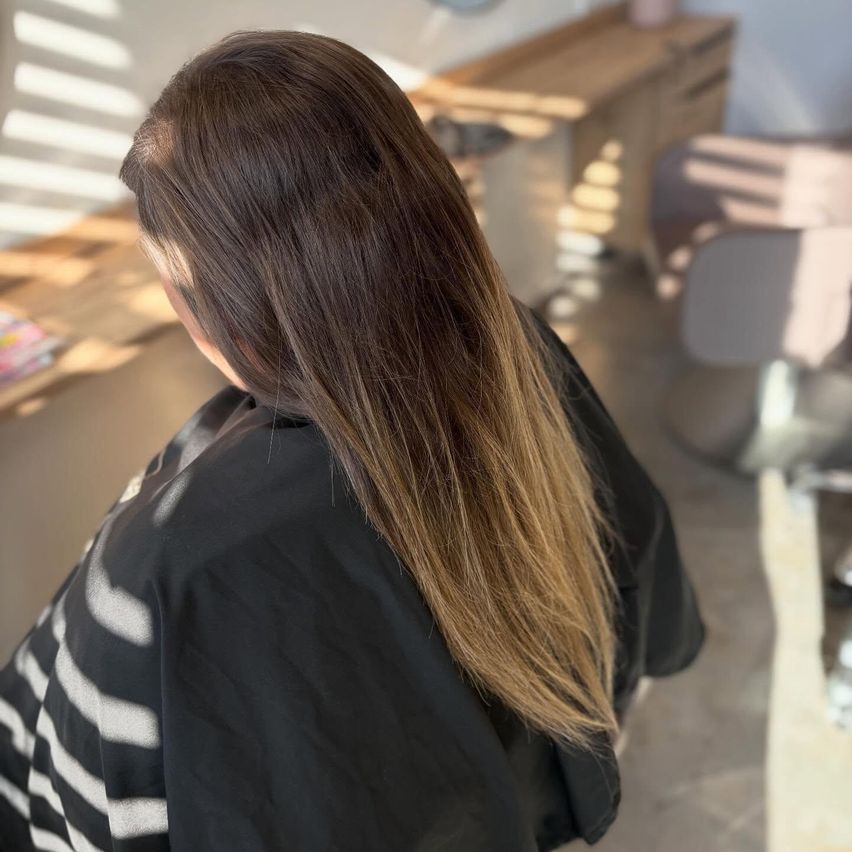 Eine Frau mit langen, glatten braunen Haaren sitzt in einem Salonstuhl und schaut nach unten. Im Hintergrund befindet sich ein Holzschreibtisch und ein Stuhl.