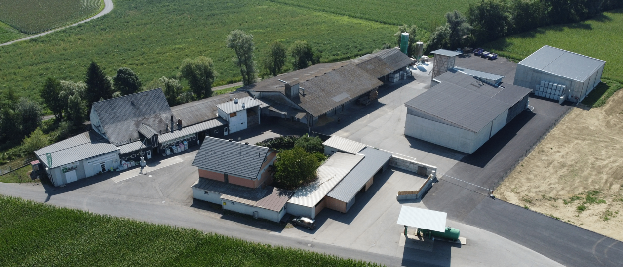 Luftaufnahme eines Bauernhofs mit mehreren Gebäuden, einschließlich eines Stalls, umgeben von grünen Feldern. Ein Auto ist auf dem Parkplatz abgestellt.