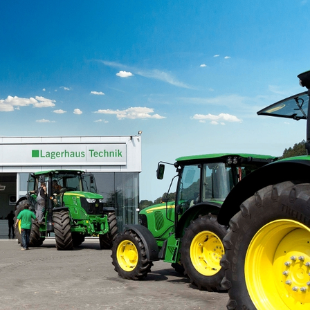 Einige John Deere Traktoren sind vor dem Lagerhaus Technik Gebäude geparkt. Ein Mann in einem grünen Shirt steht vor einem der Traktoren.