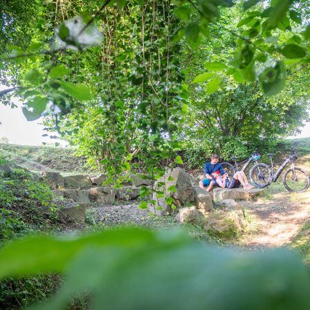 Zwei Personen ruhen sich auf einem Steinpfad mit ihren E-Bikes aus. Die Umgebung ist ein schattiger Wald mit üppigem Grün. Sie scheinen eine Pause nach einer Fahrt einzulegen.