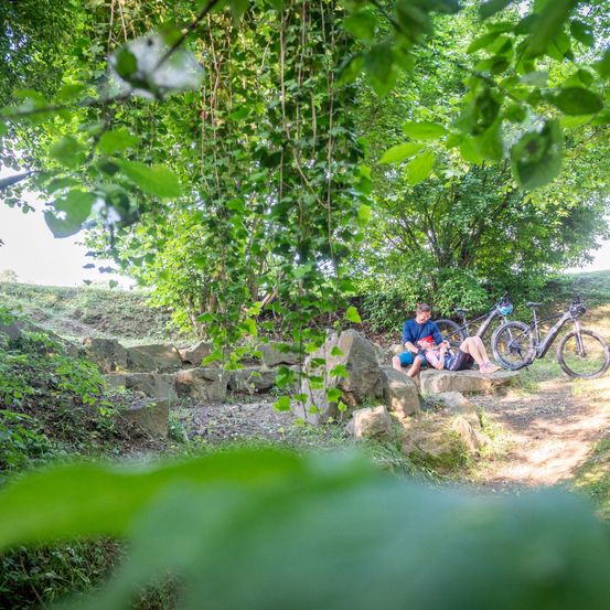 Two people rest on a stone path with their electric bikes. The setting is a shaded forest with lush greenery. They appear to be taking a break after a ride.