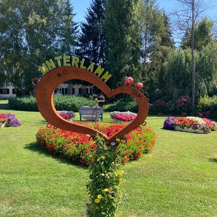 Ein Garten mit einem herzförmigen Schild, das 'Unterlamm' besagt, umgeben von bunten Blumen. Bäume und eine Bank im Hintergrund.