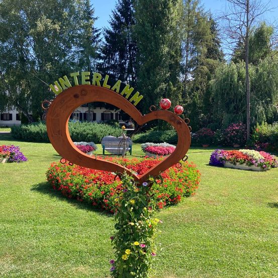 A garden with a heart-shaped sign that reads 'Unterlamm' surrounded by colorful flowers. Trees and a bench are in the background.