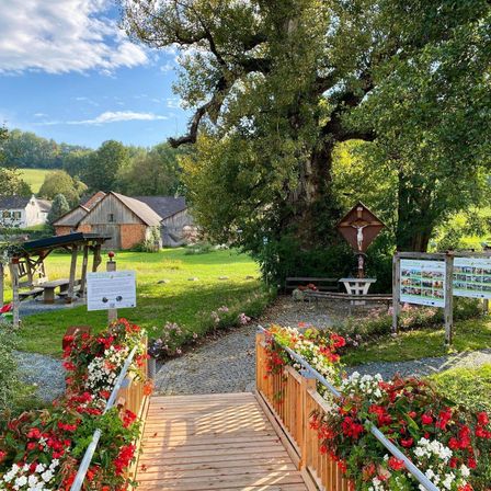 Ein ruhiger Gartenweg führt zu einer hölzernen Brücke, geschmückt mit leuchtenden roten und weißen Blumen, unter einem großen Baum mit einem Kreuz und Informationsschildern.