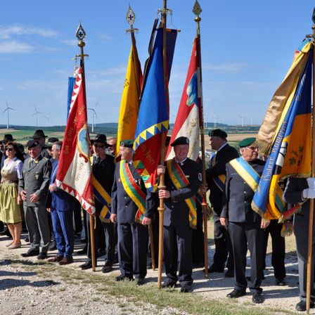 Eine Gruppe von Männern in Uniform, die Fahnen halten, möglicherweise eine feierliche Veranstaltung, steht in einer Reihe mit einigen Leuten dahinter. Der Himmel ist klar mit einigen Wolken, und es gibt Windräder in der Ferne.