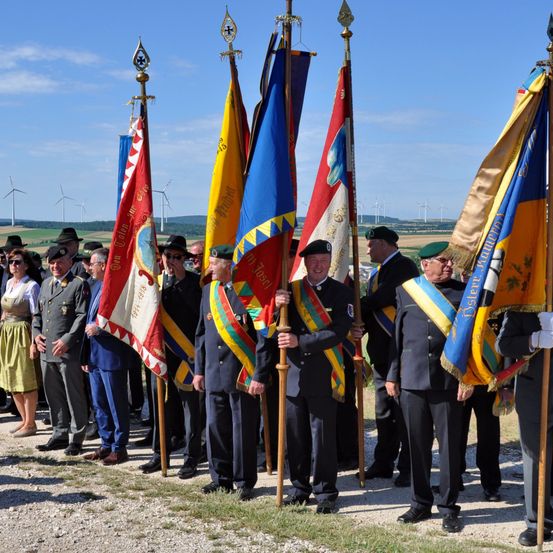 Eine Gruppe von Männern in Uniform, die Fahnen halten, möglicherweise eine feierliche Veranstaltung, steht in einer Reihe mit einigen Leuten dahinter. Der Himmel ist klar mit einigen Wolken, und es gibt Windräder in der Ferne.