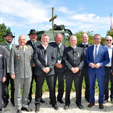 Eine Gruppe von Männern in formeller Kleidung, die vor einem Militärfahrzeug stehen, mit einem Kreuz im Hintergrund.