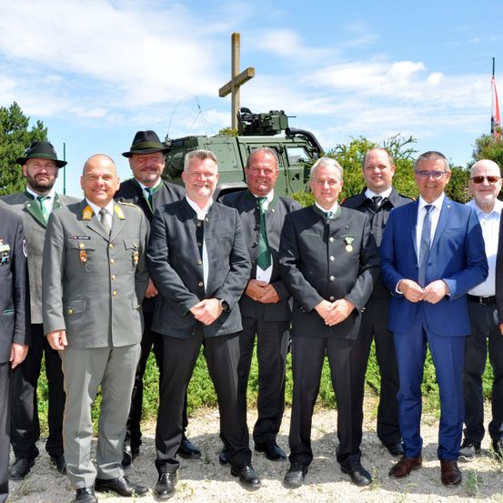 Eine Gruppe von Männern in formeller Kleidung, die vor einem Militärfahrzeug stehen, mit einem Kreuz im Hintergrund.