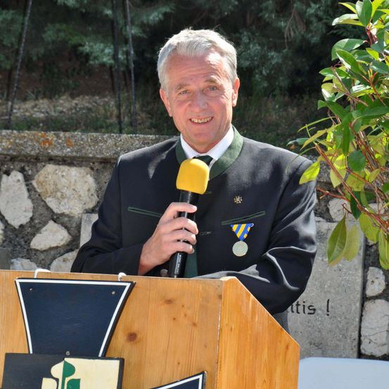 Ein lächelnder Mann in einem schwarzen Anzug hält ein Mikrofon und steht an einem Podium, mit einer grünen Schärpe und einer Medaille um den Hals. Eine Pflanze befindet sich auf der rechten Seite des Bildes.