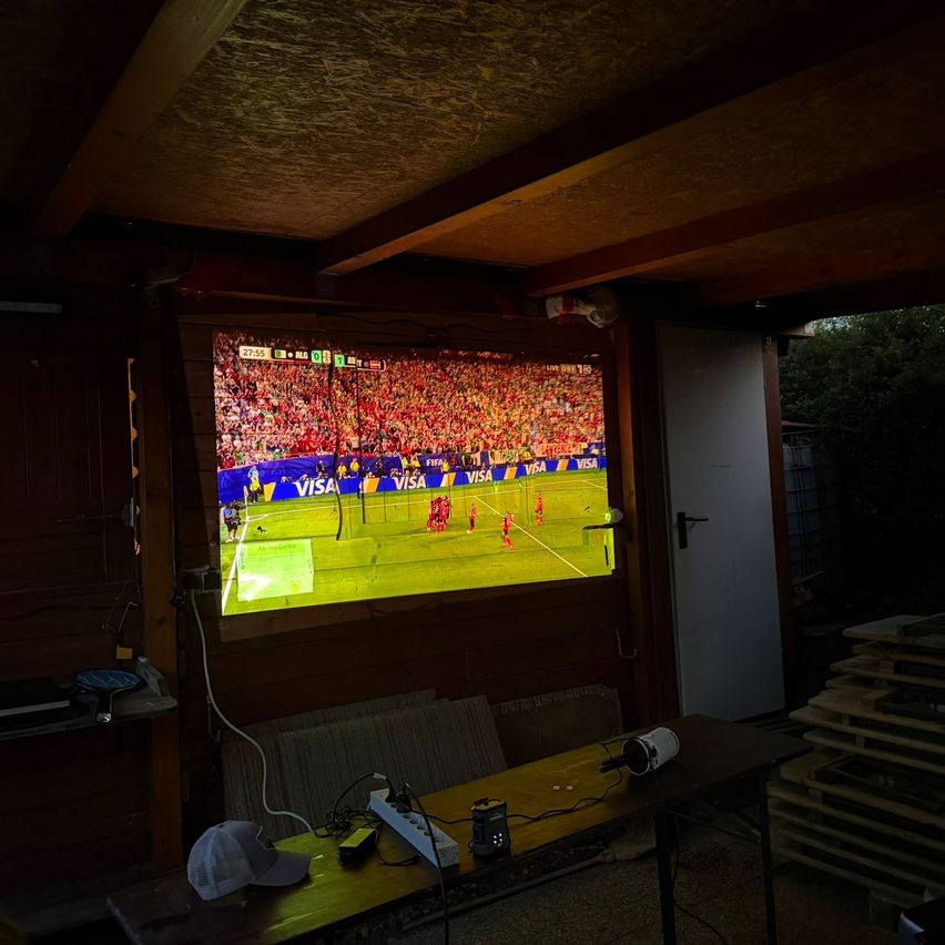 Ein großer Bildschirm in einem Raum zeigt ein Fußballspiel an. Ein Projektor steht auf einem Tisch vor dem Bildschirm, mit einem Netzteil und einem weißen Hut.