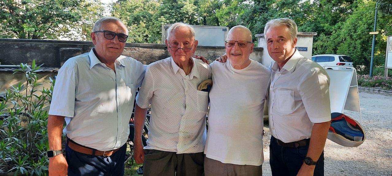 Vier ältere Männer posieren für ein Foto im Freien. Sie stehen vor einer Mauer mit Bäumen im Hintergrund. Sie alle lächeln und scheinen Freunde zu sein.