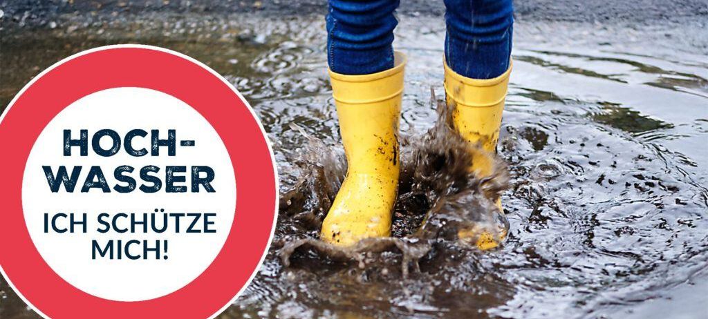 Person in gelben Gummistiefeln, die durch eine Pfütze mit dem Text 'Hochwasser schütze mich!' in deutscher Sprache im Hintergrund geht.