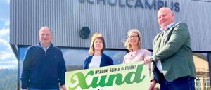 Vier Personen stehen vor einem Gebäude mit der Aufschrift Schulcampus. Sie halten ein grünes Plakat mit einer Veranstaltungsankündigung für Xund am 29. März 2026.