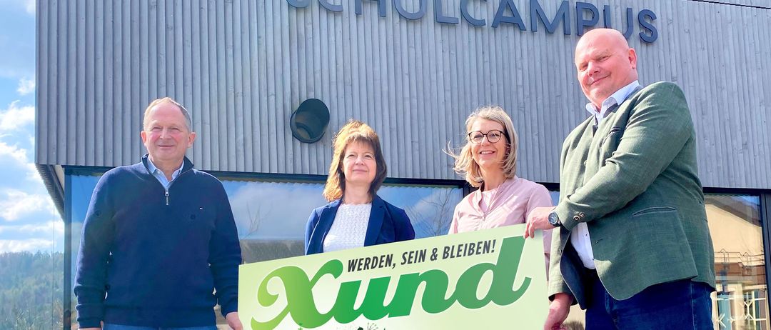 Vier Personen stehen vor einem Gebäude mit der Aufschrift Schulcampus. Sie halten ein grünes Plakat mit einer Veranstaltungsankündigung für Xund am 29. März 2026.