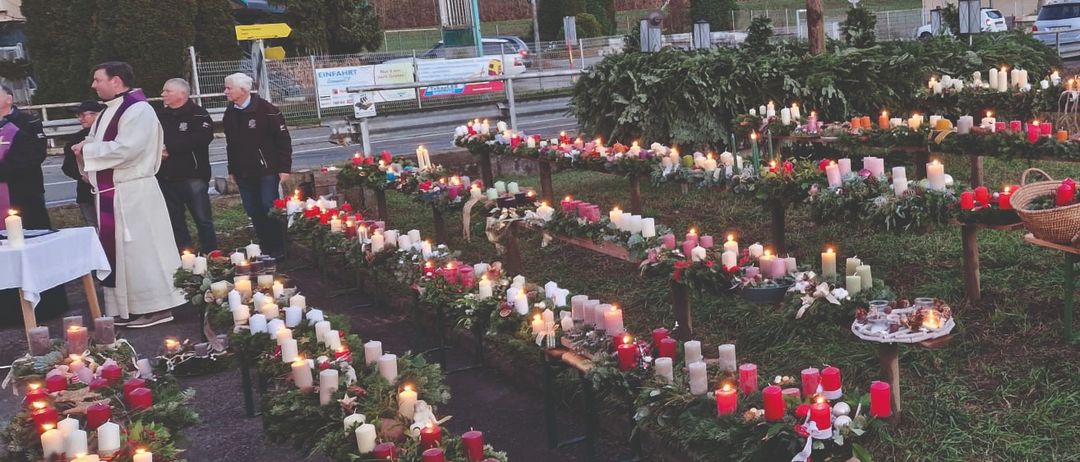 Im Freien ausgestellte zahlreiche Kerzen in Rot, Weiß und Orange, in Reihen auf einem Rasenfeld angeordnet. Ein großer Stern befindet sich in der Mitte, mit einem Haus und Fahrzeugen im Hintergrund.