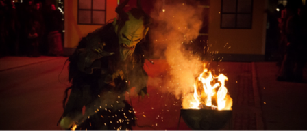 Eine dämonische Figur bläst ein Feuer an, um es aufzuflackern. Im Hintergrund beobachten ihn Menschen.