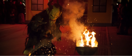 Eine Person in einer gehörnten Maske führt einen Ritus vor einem brennenden Feuer durch, wobei Rauch aufsteigt und Menschen im Hintergrund zuschauen.