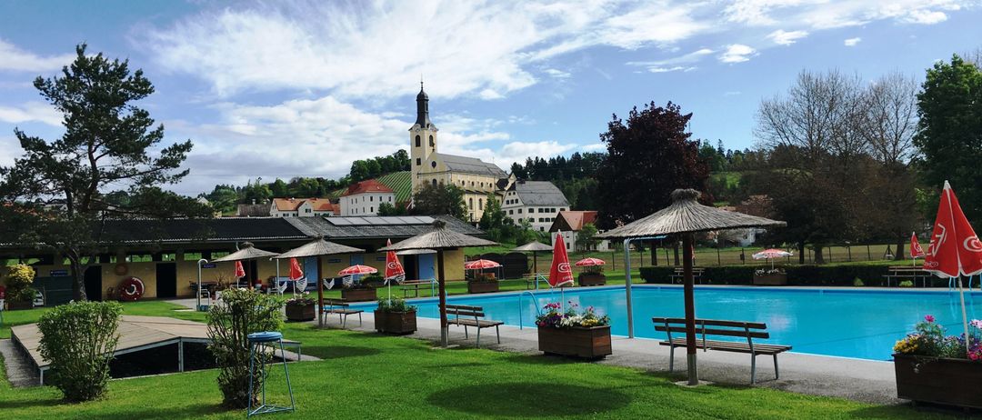 Ein großer Außenpool, umgeben von einem üppigen grünen Rasen, mit Bänken und Sonnenschirmen, unter einem teilweise bewölkten Himmel, mit einer Kirche im Hintergrund.