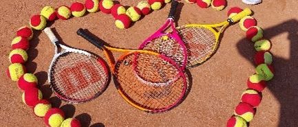 Vier junge Mädchen sitzen um ein Herz aus Tennisbällen und Schlägern herum und lächeln für die Kamera auf einem Tennisplatz.