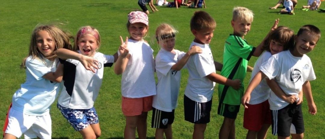 Eine Gruppe von Kindern in Sportkleidung steht auf einem Rasenfeld, einige lächeln, andere sind im Hintergrund mit verschiedenen Aktivitäten beschäftigt.