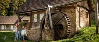 Three people stand in front of an old brick building with a large wheel. The building is surrounded by grass, and there are trees in the background.