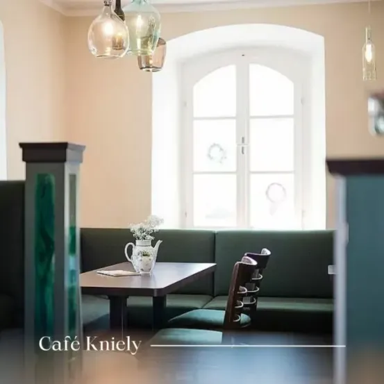 Eine gemütliche Caféecke mit Tisch und Stühlen. Auf dem Tisch steht eine Vase mit Blumen. Eine Bank steht neben dem Fenster. Lampen hängen von der Decke.