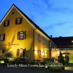 Das Kniely Haus Leutschach zeigt ein gelbes Gebäude mit mehreren Fenstern. Zwei Personen sitzen draußen und genießen den Abend. Der Name Kniely Haus Leutschach steht unten.