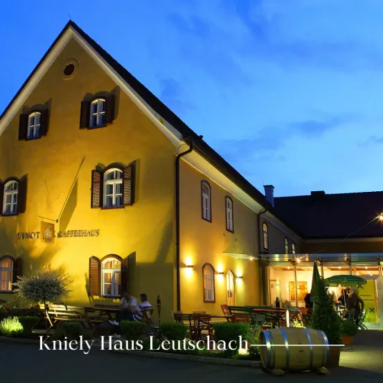 Das Kniely Haus Leutschach zeigt ein gelbes Gebäude mit mehreren Fenstern. Zwei Personen sitzen draußen und genießen den Abend. Der Name Kniely Haus Leutschach steht unten.