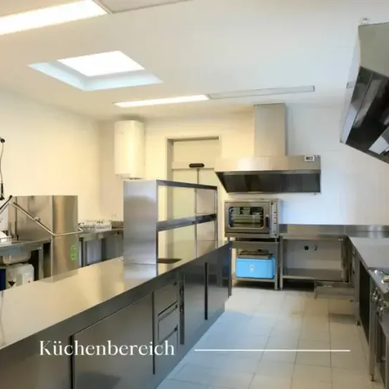 Ein Küchenbereich mit Edelstahlschränken und -theken. Ein Ofen, eine Mikrowelle und ein Kühlschrank stehen auf der Arbeitsplatte. Eine Spüle mit Wasserhahn befindet sich neben dem Kühlschrank. Eine Dunstabzugshaube ist an der Wand montiert. Die Decke ist weiß.