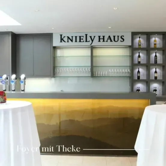 Eine Bartheke mit der Aufschrift 'KnieLy HAUS'. Zwei Tische mit weißen Tischdecken stehen auf beiden Seiten. Zwei Flaschen stehen auf der Barthekenplatte.