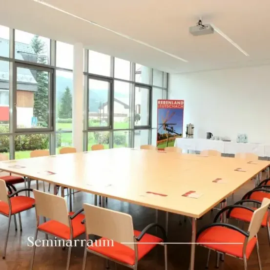 Ein Konferenzraum mit großen Fenstern, einem langen Holztisch und orangefarbenen Stühlen. Eine Projektor ist an der Decke montiert. Ein Schild ist an der rechten Wand angebracht.