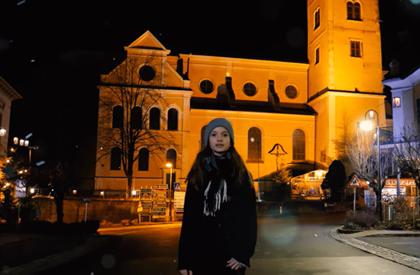Eine junge Frau steht nachts vor einer Kirche, beleuchtet von Straßenlaternen.