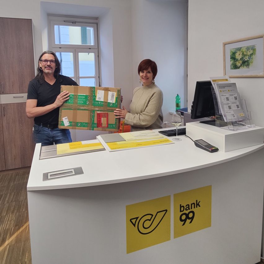 A man and woman hold a box in front of a counter with Bank 99 signage, a computer, and a glass of wine on a desk. Behind them is a window and a picture frame.