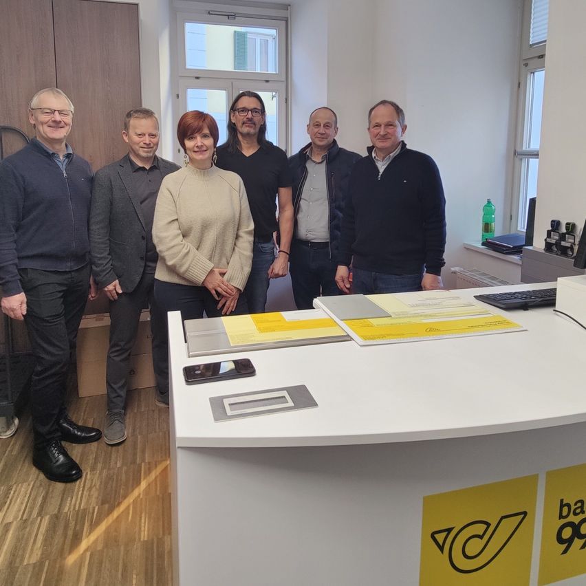 Five people stand in an office in front of a desk with a yellow sign. Papers and a phone are on the desk.