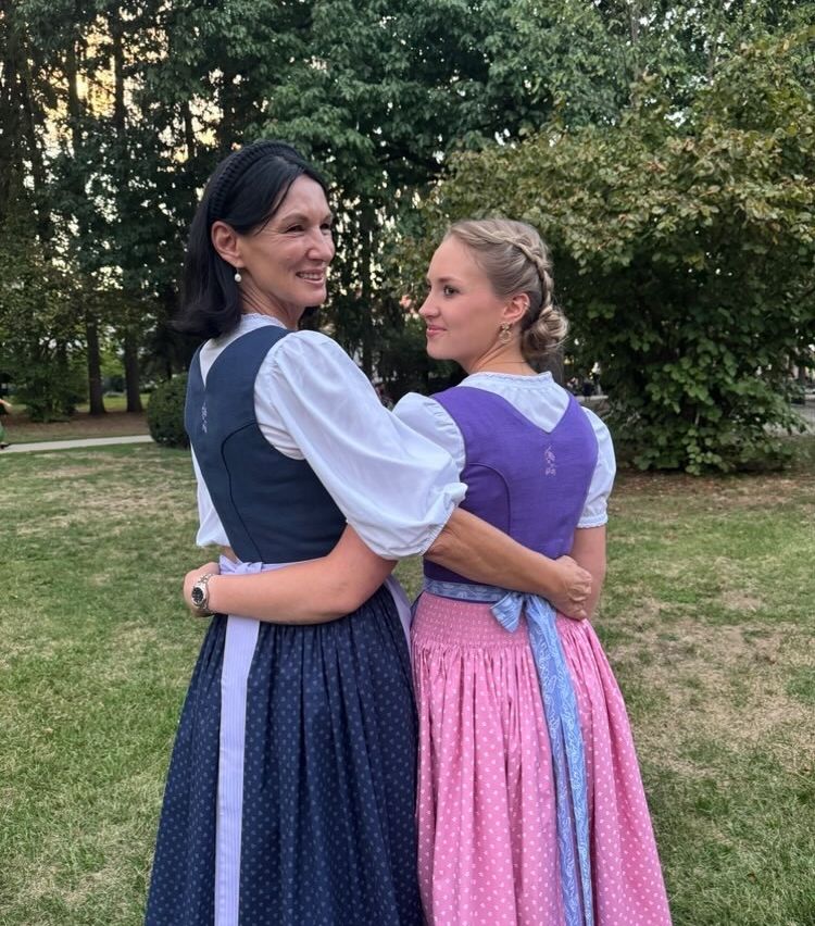 Zwei Frauen in traditioneller Kleidung stehen eng beieinander auf einem Rasen und lächeln in die Kamera.