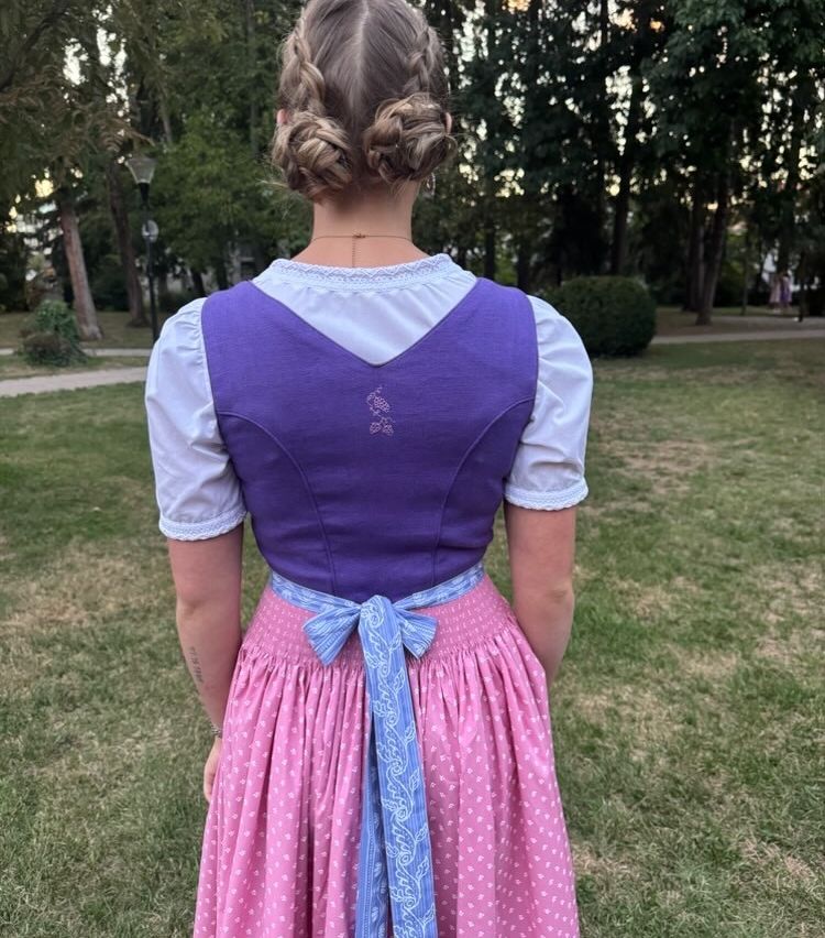 Eine Frau mit geflochtenen Haaren steht in einem Park und trägt ein lila und weißes Oberteil mit einem blauen Gürtel um die Taille und einen rosa Rock.