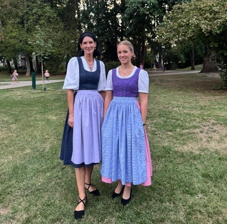 Zwei Frauen in traditionellen Kleidern stehen auf einem Rasen und lächeln für ein Foto. Bäume und ein Weg sind im Hintergrund.