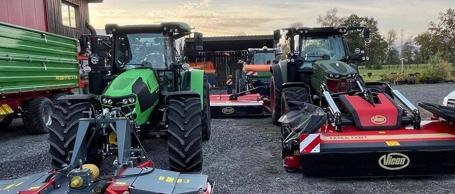 Mehrere Traktoren sind vor einem Scheunen bei Sonnenuntergang geparkt. Einer hat eine rote Mähmaschine angeschlossen.