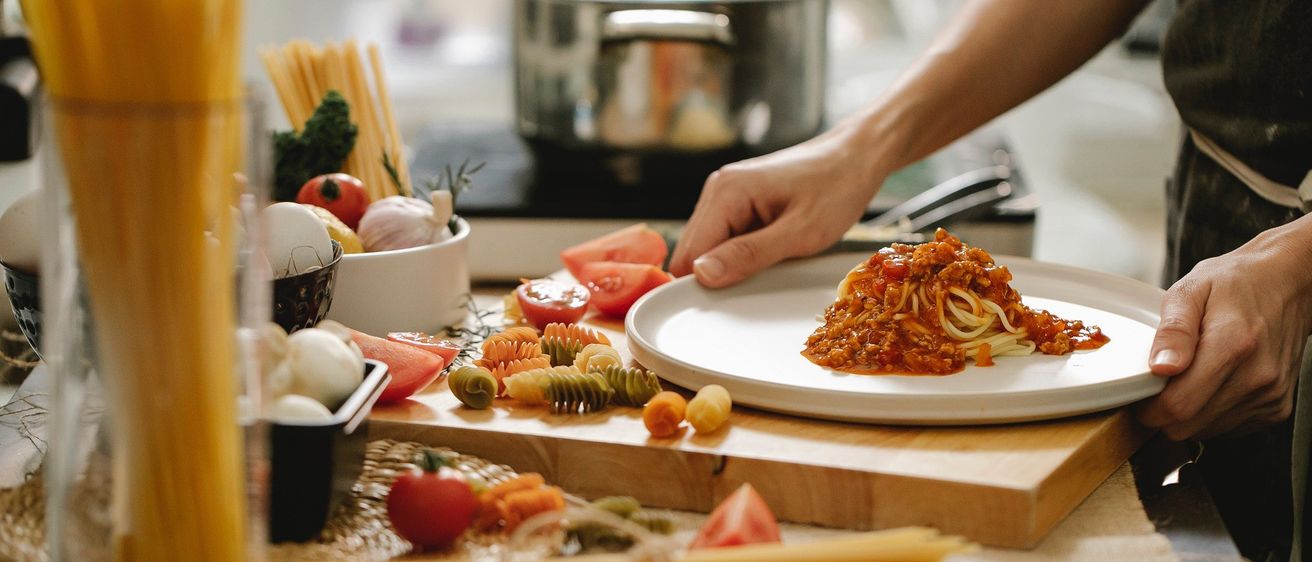 Eine Person serviert Spaghetti mit Fleischsauce auf einem Küchentisch, umgeben von verschiedenen Zutaten.