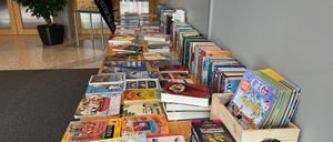 Ein langer Tisch, gefüllt mit verschiedenen Kinderbüchern, einige stehen aufrecht und andere liegen herum. Rechts steht ein Holzkasten mit Zeitschriften und Büchern. Ein Banner mit dem Wort 'Flohmarkt' steht hinter dem Tisch. Links steht ein Topf mit einer Pflanze.