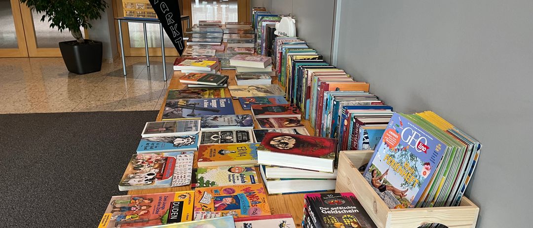 Ein langer Tisch, gefüllt mit verschiedenen Kinderbüchern, einige stehen aufrecht und andere liegen herum. Rechts steht ein Holzkasten mit Zeitschriften und Büchern. Ein Banner mit dem Wort 'Flohmarkt' steht hinter dem Tisch. Links steht ein Topf mit einer Pflanze.