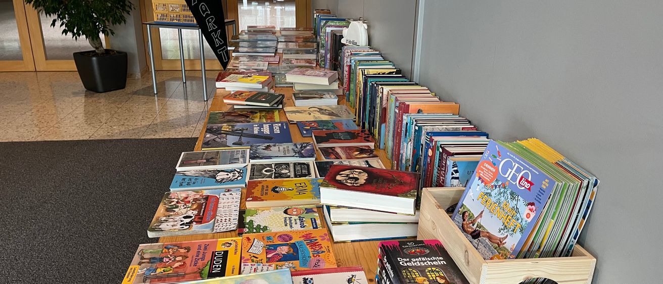 Ein langer Tisch, gefüllt mit verschiedenen Kinderbüchern, einige stehen aufrecht und andere liegen herum. Rechts steht ein Holzkasten mit Zeitschriften und Büchern. Ein Banner mit dem Wort 'Flohmarkt' steht hinter dem Tisch. Links steht ein Topf mit einer Pflanze.