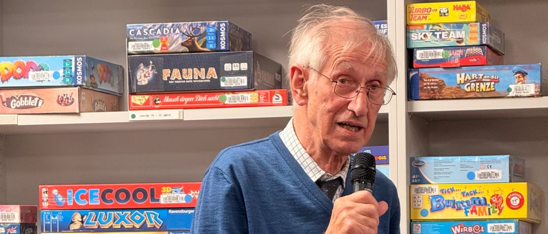 Ein älterer Mann in einem blauen Pullover spricht in ein Mikrofon in einem Raum voller Brettspiele an Regalen.