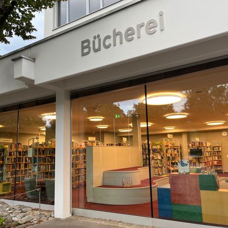 Das Äußere einer Buchhandlung mit dem Wort Bucherei über den Glastüren. Im Inneren befinden sich mit Büchern gefüllte Regale, eine Treppe und farbenfrohe Fächer.