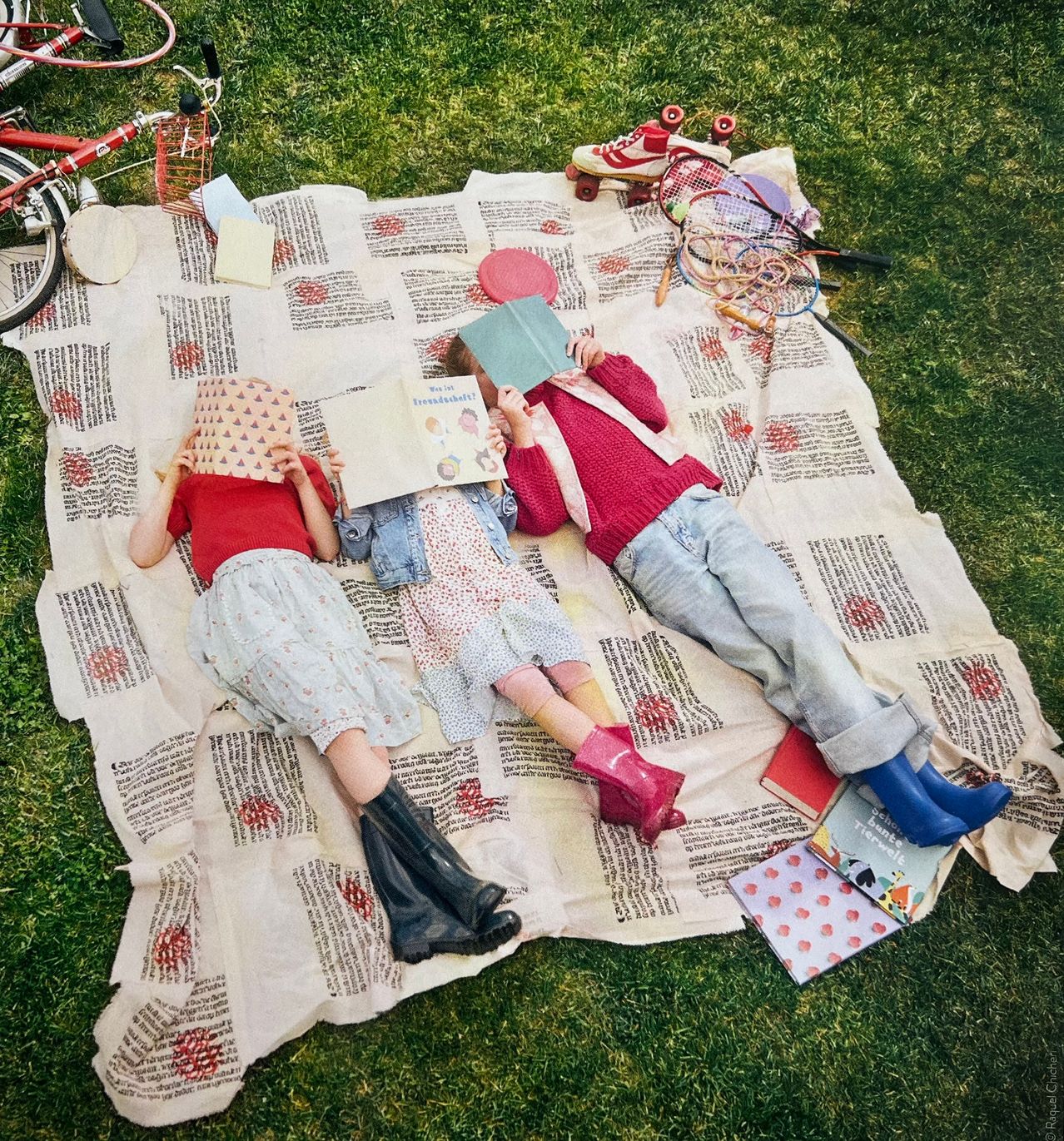 Three children lie on a blanket reading books. A bicycle, a basket, and roller skates are around them. A small toy is on the blanket.